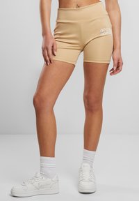 Beige shorts met hoge taille gemaakt van rekbaar materiaal met een gladde textuur, met witte "K" letters aan de zijkant. Gecombineerd met witte sneakers.