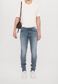Muž v světlých modrých skinny jeans, bílém polo tričku, bílých teniskách a malém tmavě hnědém křížovém vaku na pozadí bez detailů.