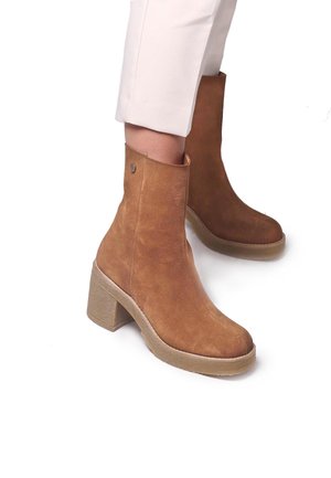 Braune Wildleder-Stiefeletten mit einem hohen Blockabsatz, abgerundeter Zehenpartie und einem kleinen metallischen Akzent am oberen Rand. Weiche Textur und minimalistisches Design.