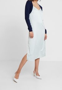 Satin klänning i ljusblått med djup V-ringning och marinblå långärmar. Åtföljd av vita högklackade skor, slim fit, knälång.