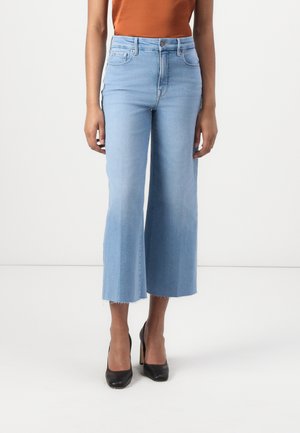 Kvinde iført lyseblå korte flare-jeans med rå kant, kombineret med sorte højhælede sko og en orange ærmeløs top.