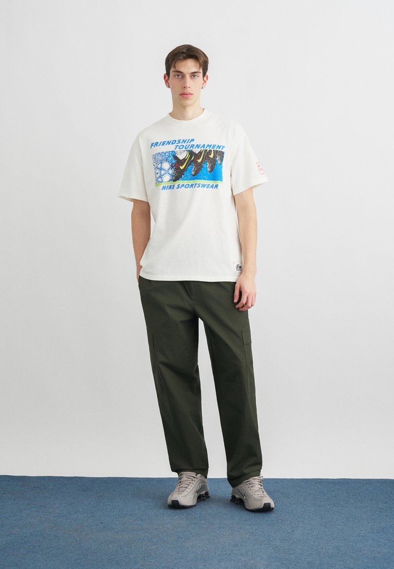 Junger Mann steht mit den Händen in den Taschen, trägt ein weißes Nike-Grafik-T-Shirt, olivgrüne Cargohosen und beige Turnschuhe auf blauem Teppich.