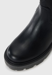 Close-up van een zwarte leren enkelboot met een ronde neus en een gestructureerde rubberen zool op een witte achtergrond.