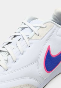 Λευκό αθλητικό παπούτσι με μπλε και ροζ λογότυπο Nike, διαθέτοντας υφασμάτινες επιφάνειες, ανοιχτό γκρι κορδόνια και λεία, συνθετική επένδυση.