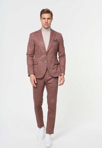Traje para hombre en tela texturizada color burdeos, con solapa de pico, cierre de un solo botón y pantalones slim-fit a juego. Zapatillas blancas complementan el look.