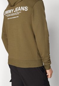 Muž v olivově zelené mikině s bílým logem "Tommy Jeans" a názvy měst na zádech, v kombinaci s černými kalhotami.