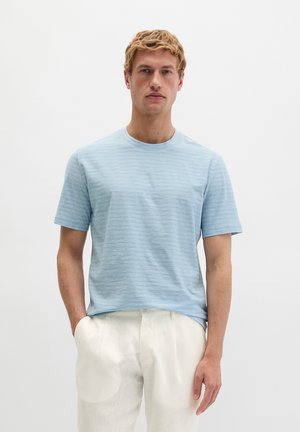 Man wearing a light blue short-sleeve t-shirt and white pants standing against a plain white background.