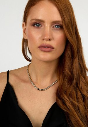 Halskette mit abwechselnd klaren und schwarzen quadratischen sowie rechteckigen Steinen, die in einer goldfarbenen Kette eingefasst sind, getragen mit einem schwarzen Oberteil.