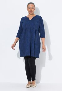 Donna che indossa una tunica blu a motivi con maniche a tre quarti, leggings neri, scarpe piatte dorate e orecchini a cerchio dorati, in piedi davanti a uno sfondo bianco.