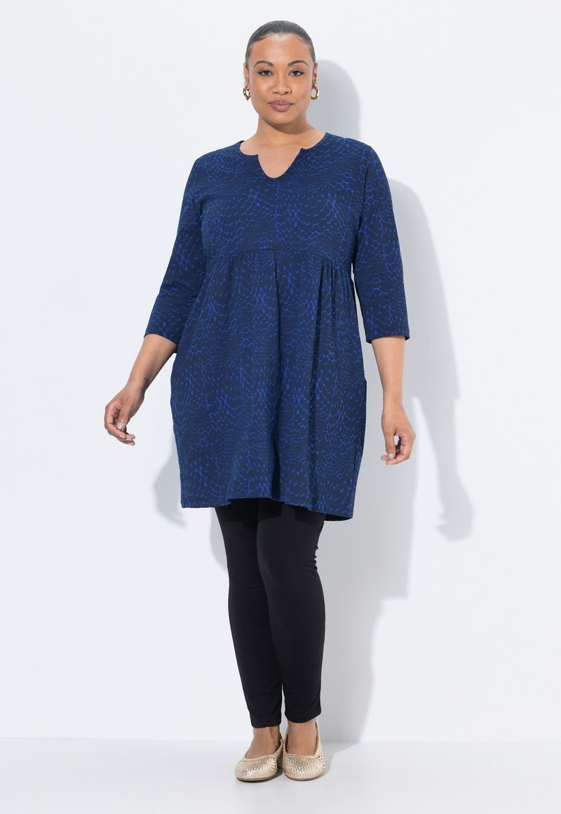 Donna che indossa una tunica blu a motivi con maniche a tre quarti, leggings neri, scarpe piatte dorate e orecchini a cerchio dorati, in piedi davanti a uno sfondo bianco.