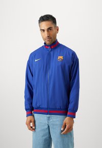 Modrá športová bunda s vertikálnymi pruhmi, zipsom, vysokým golierom, červenými a modrými akcentmi, logom Nike a emblémom Barcelony na hrudi.