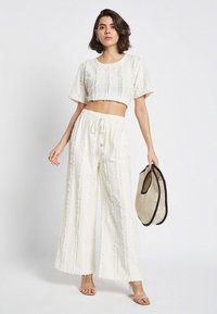 Weißes Zweiteiler-Outfit mit strukturierten, ausgefransten Details. Kurzes Oberteil kombiniert mit weit geschnittenen Hosen. Accessories umfassen eine geflochtene Tasche und transparente Absätze.