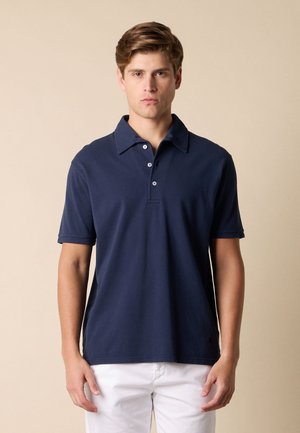 Młody mężczyzna z krótkimi brązowymi włosami, ubrany w granatową koszulkę polo z krótkim rękawem i białe spodnie na gładkim beżowym tle.