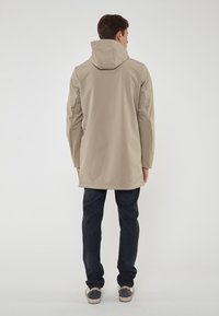 Cappotto impermeabile beige con cappuccio, maniche lunghe e vestibilità comoda, abbinato a jeans scuri e scarpe casual. Texture liscia, design minimale.