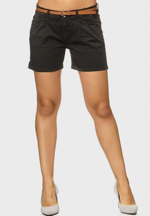 Femme portant un short noir avec ceinture et des chaussures à talons hauts gris clair, debout devant un fond clair uni.