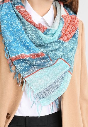 Femme portant une écharpe à motifs multiples dans des nuances de bleu, rouge et sarcelle, sur une chemise blanche et un manteau couleur camel.