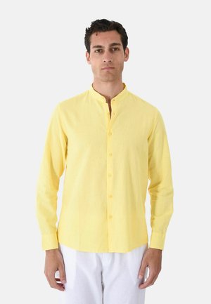 Uomo che indossa una camicia gialla a maniche lunghe con colletto alla coreana e pantaloni bianchi, in piedi davanti a uno sfondo chiaro uniforme.