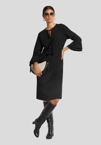 Schwarzes knielanges Kleid mit einem gebundenen Ausschnitt, ausgestellten Ärmeln und glatter Textur. Kombiniert mit knielangen schwarzen Stiefeln und einer Halbmond-Handtasche.