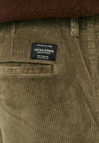 Olive green corduroy pants with vertical ribbing, featuring a black label with white text reading "JACK & JONES PANTS STUDIO."