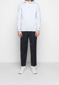 Felpa grigia chiara con tasca a marsupio e logo su gruccia, abbinata a pantaloni blu scuro e sneaker bianche con dettagli blu.