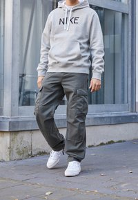Šedá mikina s kapucí s černým nápisem "NIKE", doplněná zelenými cargo kalhotami s bočními kapsami a bílými teniskami na dlážděném povrchu.