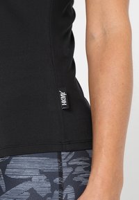 Svart atletisk linne med smal passform. Har en liten varumärkesetikett på sidan. Bärs med mönstrade svarta leggings med vertikala ränder.