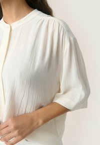 Šviesiai kreminė bluzelė su švelnia tekstūra, pusilgiais rankovėmis ir mandarinų apykakle; turi subtilias vertikalias raukšles palei kūną.