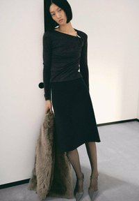 Top negru cu maneci lungi și un decolteu unic, asociat cu o fustă neagră de lungime până la genunchi. Modelul ține o haină din blană sintetică de culoare bej deschis. Pantofi cu toc gri.