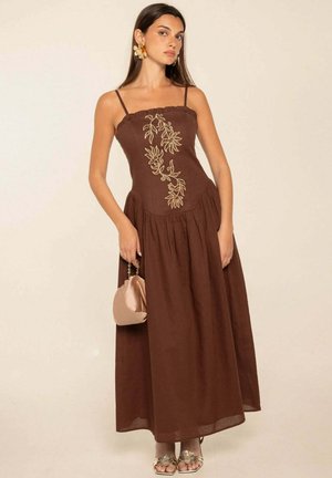 Mujer con un vestido sin mangas marrón con bordados florales, sosteniendo un pequeño bolso beige y llevando pendientes florales dorados y sandalias.