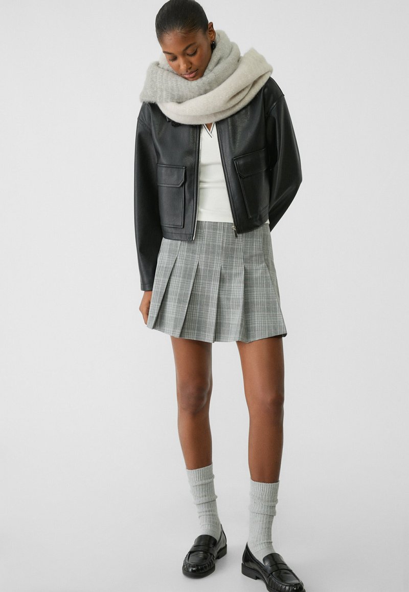 Veste en cuir noire courte, haut blanc, jupe plissée grise à carreaux, chaussettes tricotées grises à motifs et mocassins noirs.