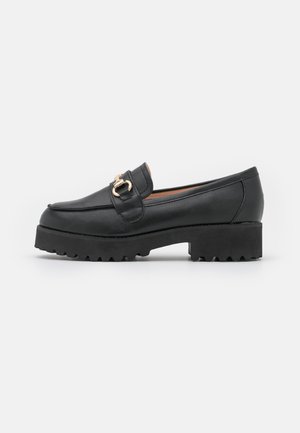 Mocassins en cuir noir avec une semelle en caoutchouc épaisse, dotés d'une boucle dorée à l'avant et d'une texture lisse et élégante.