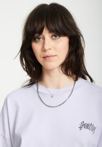 Licht paarse katoen T-shirt met een relaxte pasvorm, voorzien van een zwart logo op de linkerborst. Twee zilveren kettingen zichtbaar, waarvan één met een sterrenhanger.