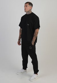 Černé oversized tričko s vyšívaným logem, spárované s černými kalhotami typu cargo s vyšívaným detailem, doplněné bílými teniskami.