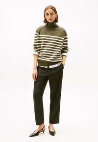 Woman wearing olive green and white striped turtleneck sweater, white shirt, dark pinstripe trousers, and black slingback heels standing against white background.
