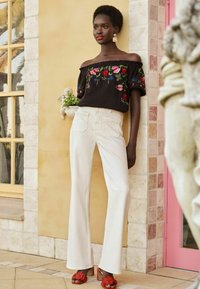 Off-the-shoulder black top with floral embroidery, paired with white flared pants. Red heeled sandals with floral accents. Smooth textures.