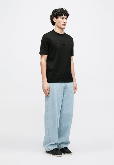 Μαύρο βαμβακερό t-shirt με ανάγλυφο λογότυπο "Emporio Armani", σε συνδυασμό με ανοιχτού μπλε baggy τζιν και μαύρα αθλητικά παπούτσια, στέκεται σε ουδέτερο φόντο.