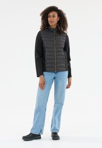 Svart vadderad väst med hög krage och guld zip; långa svarta ärmar; tillsammans med ljusblå jeans och svarta skor.
