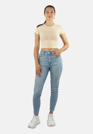 Helles beige Crop-Top, hochgeschnittene blaue Jeans und weiße Sneakers. Das Outfit zeichnet sich durch ein enganliegendes Design mit kontrastierenden Farben aus.
