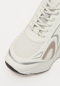 Hvide sneakers med mesh- og syntetiske materialer, struktureret overflade, grå accenter og flade snørebånd på en rund tåform. Sort sål.