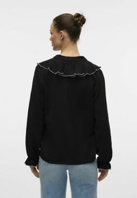 Sort langærmet top med en volangkrave med hvid kant. Stofoverfladen har en struktureret udseende. Sammen med lyseblå jeans.