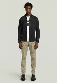 Camicia in denim nera a bottoni sopra una t-shirt grafica bianca, abbinata a pantaloni cargo beige e stivali alla caviglia neri, con molteplici tasche.