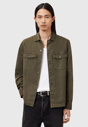 Olive green cotton jacket featuring a pointed collar, two chest pockets with button closures, and a relaxed fit, worn over a white shirt.