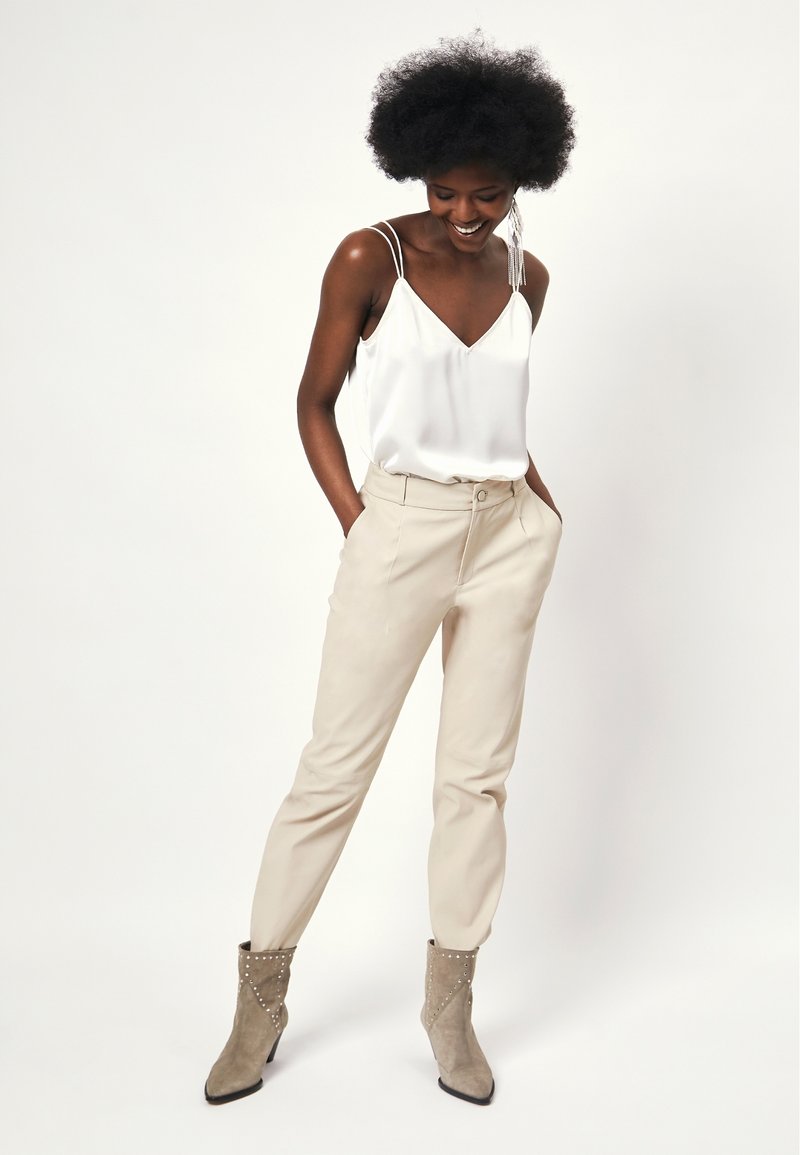 Witte satin camisole met dunne bandjes, gecombineerd met beige tapered broeken. Model draagt grijze enkellaarzen met studs.