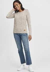 Strickpullover in hellem Braun mit strukturiertem Muster und siden Knöpfen, kombiniert mit blauen Jeans im geraden Schnitt und weißen Turnschuhen.