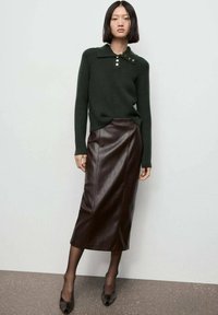 Mujer joven vestida con un suéter de canalé verde oscuro con cuello abotonado y una falda midi de cuero marrón, con unas medias negras y tacones negros, de pie.