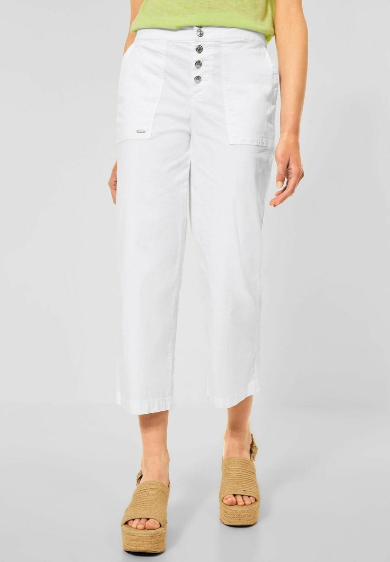 Pantalon blanc court taille haute avec quatre boutons sur le devant et grandes poches plaquées, porté avec des sandales à plateforme beiges et un haut vert.