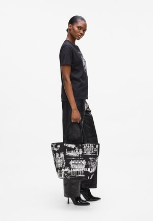 Tote bag with black fabric, featuring white architectural sketches, double handles, and a printed logo. Model wears a black shirt and shiny pants.