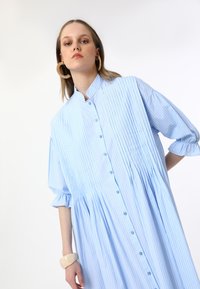 Robe surdimensionnée à rayures bleu clair, avec des manches courtes, un devant à boutons et des détails plissés. Elle présente un col haut et une taille froncée.