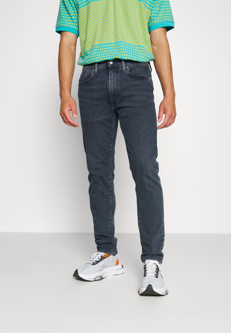 Man som bär mörka slimfit-jeans, en grön och gul mönstrad polo skjorta, och vita sneakers med orangea detaljer, stående mot en neutral bakgrund.