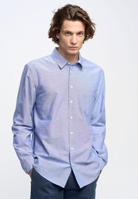 Camisa de botones azul claro con un bolsillo, de tela de algodón, de mangas largas y un diseño de cuello clásico. Llevada con pantalones oscuros.